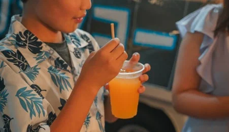 夏らしい柄のシャツを着た子供が、ストローを使ってオレンジ色の飲み物を飲んでいるクローズアップ