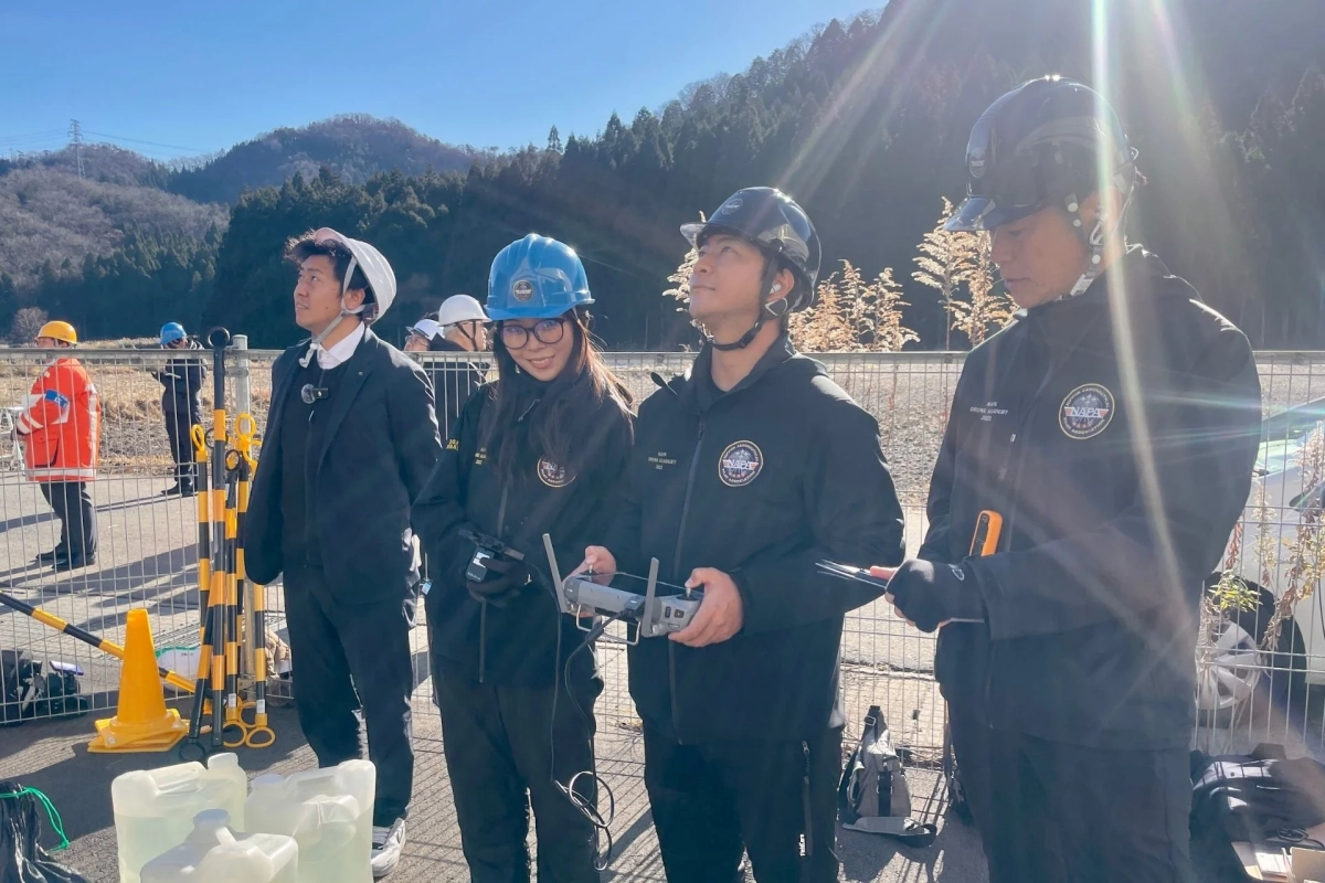 晴れた屋外で、ヘルメットを着用した人々がドローンの操縦訓練を行っています。