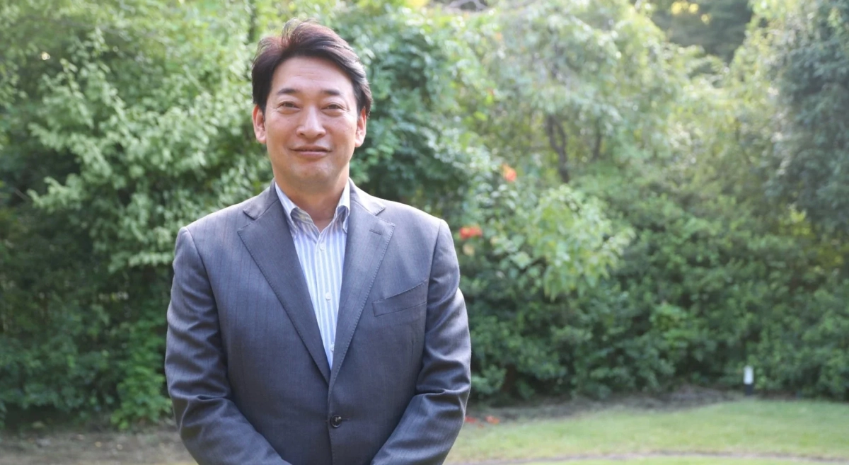 緑豊かな屋外を背景に、グレーのスーツとストライプシャツを着用したアジア人男性がカメラに向かって穏やかに微笑んでいます