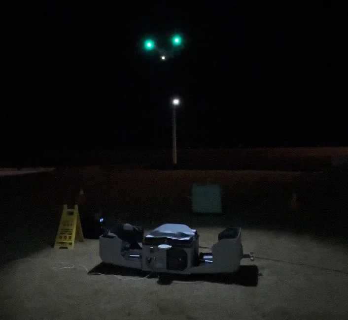 夜の屋外で撮影された画像で、上空には二つの緑色の光が浮かび、地上には機器と黄色い警告サイン、そして遠くに街灯が見える