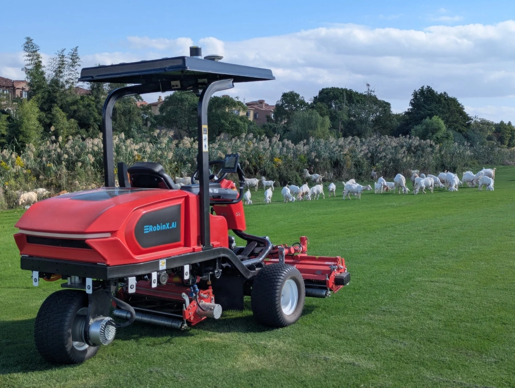 青空の下、広々とした緑の牧草地に赤い自動芝刈り機が置かれ、その奥では白いヤギの群れが草を食んでいます。