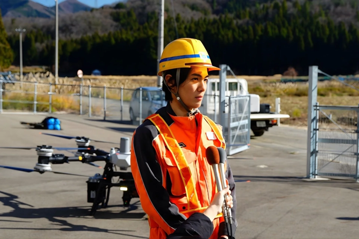 黄色のヘルメットとオレンジ色の安全ベストを着用した人物が屋外でインタビューを受けている様子。