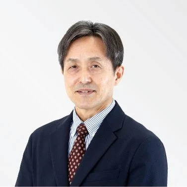 白い背景の前で、ダークスーツとネクタイを着用した中高年の男性が穏やかな笑顔を見せているポートレート写真です。