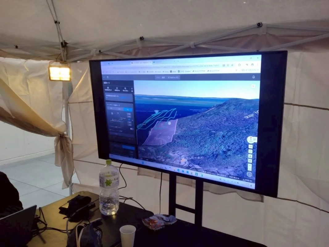 テント内に設置された大型モニターに、山岳地帯の3D地図やドローン飛行ルートらしきものが表示されています