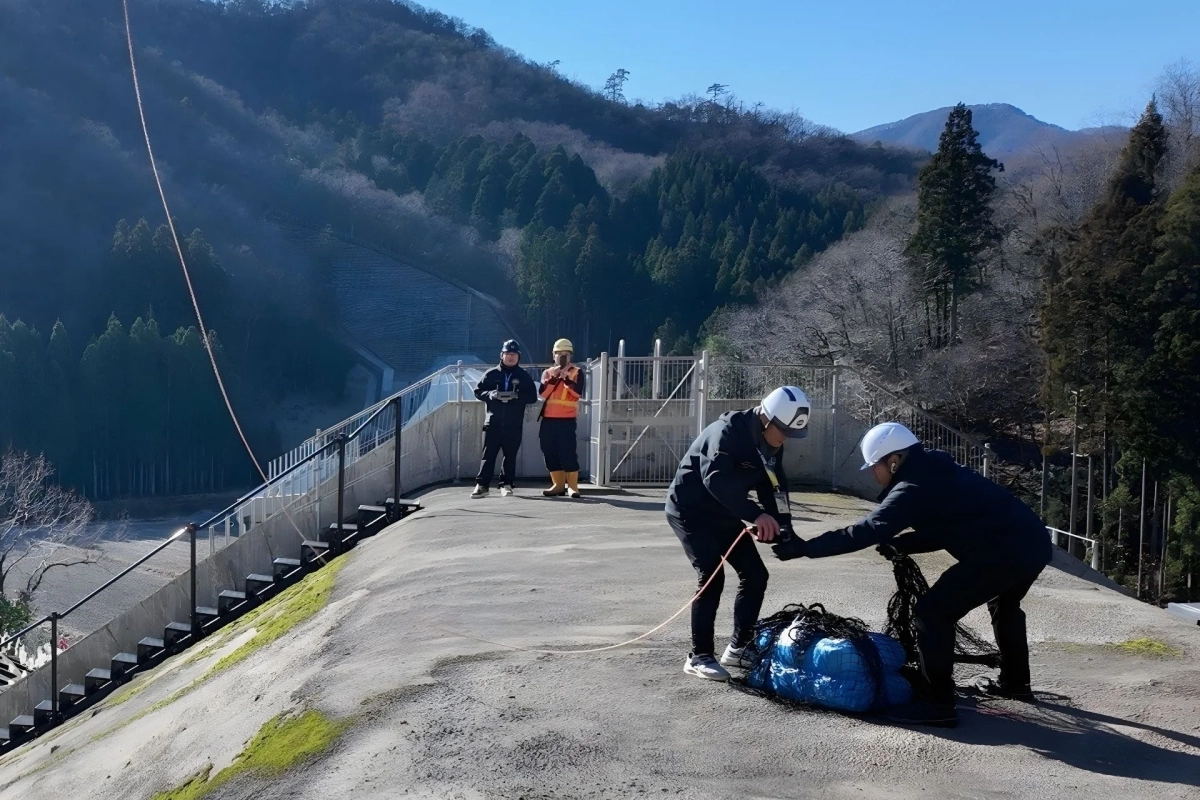 山間の場所で、複数の作業員がドローンを用いた測量準備や資材の設置作業を行っています。