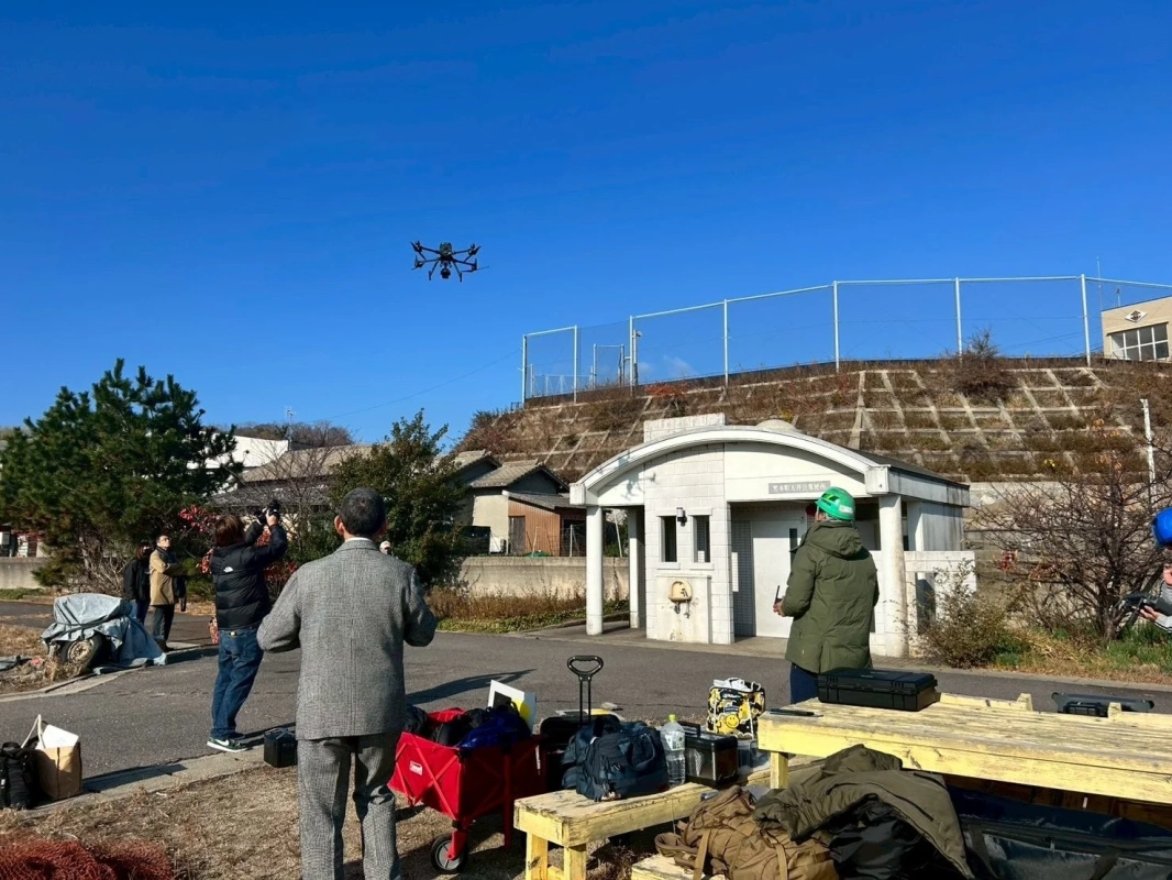 青い空の下、屋外でドローンが飛行している様子を複数の人々が見上げています