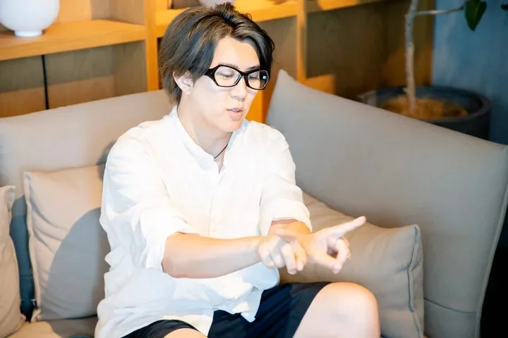 眼鏡をかけた男性が白いシャツを着てソファに座り、両手でジェスチャーを交えながら何かを説明している場面