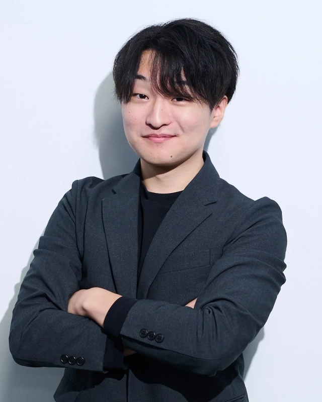 白い背景の前で、ダークグレーのスーツジャケットを着用し、腕を組んで微笑む若いアジア人男性のポートレートです。