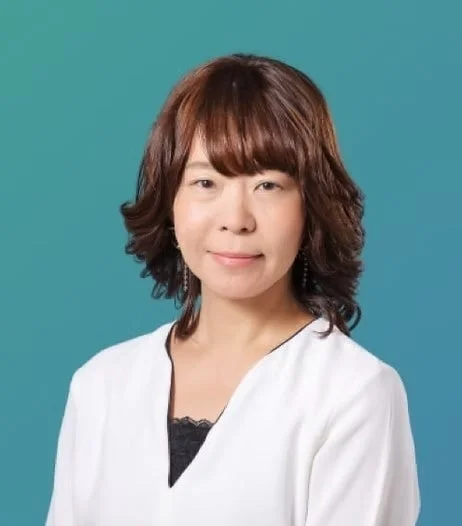 青緑色の背景を背に、白いトップスを着たアジア系の女性が穏やかに微笑んでいるポートレート写真です。