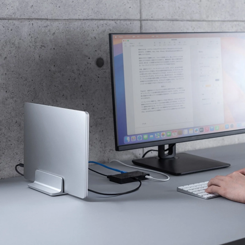 デスク上で縦置きスタンドに立てられたMacBookと外部モニターが接続され、ユーザーがキーボードを操作している