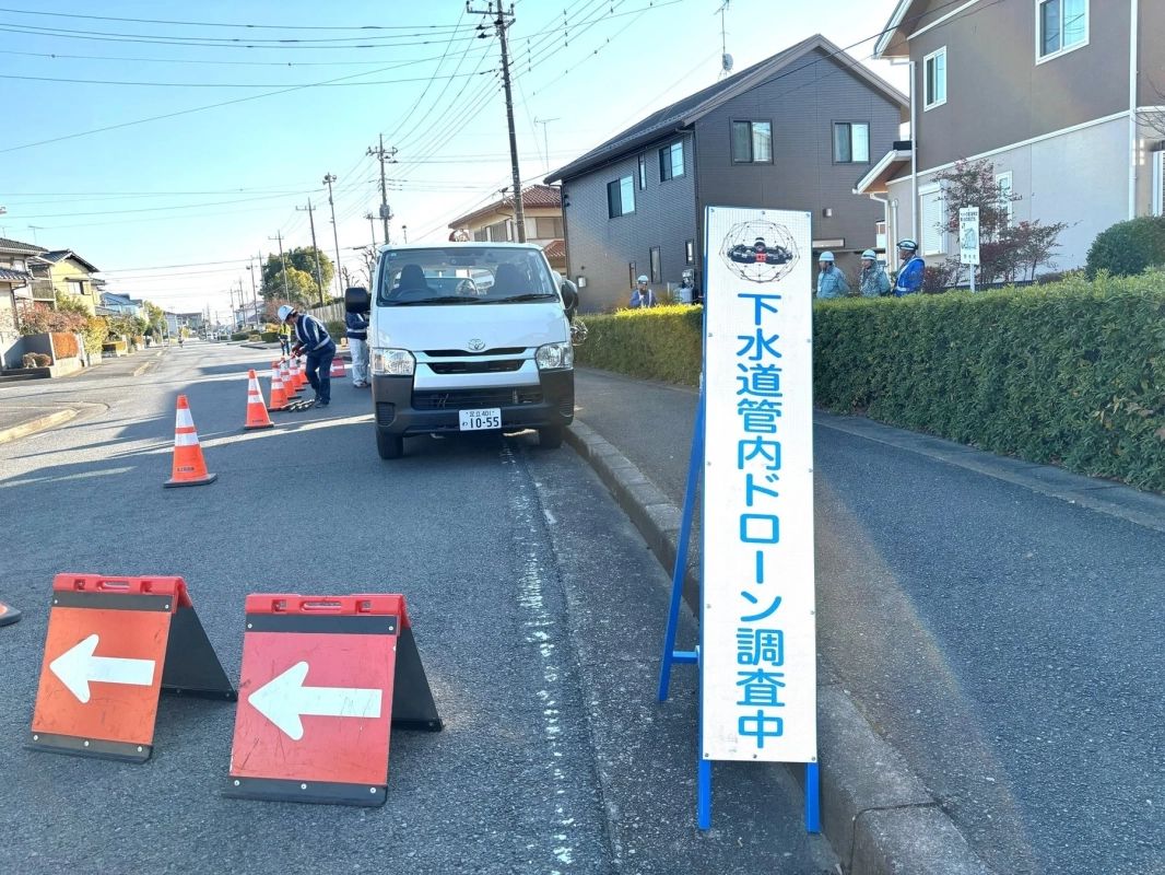 住宅街の道路で下水道管内のドローン調査が行われている風景。作業員が交通コーンや矢印看板を設置し、安全に作業を進めている様子が捉えられています。