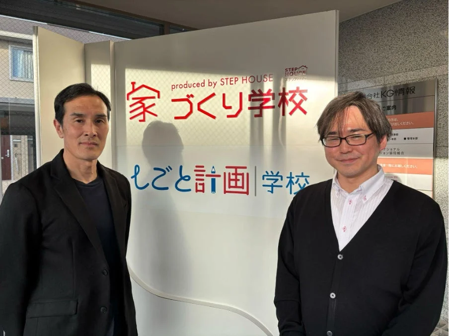 オフィスのような場所で、2人の男性が「家づくり学校」と「しごとと計画|学校」と書かれた看板の前に立っている。
