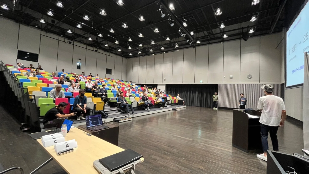 カラフルな座席が並ぶ講堂で、多くの聴衆がプレゼンテーションを聞いている様子