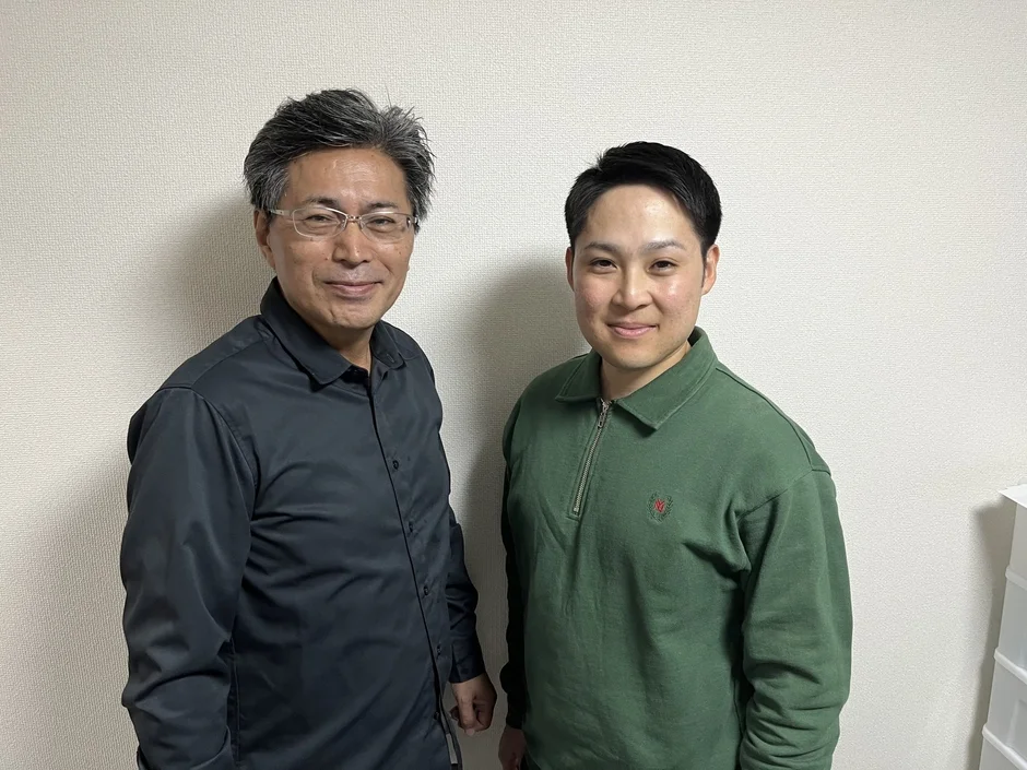 白い壁を背景に、二人の男性が並んで笑顔で立っている写真