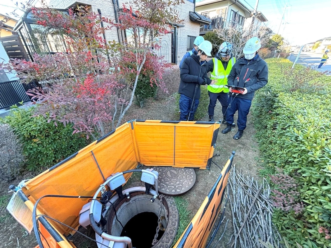 住宅街で数人の作業員がマンホールの点検を行っている様子です。オレンジ色の安全柵が設置され、特殊な機材を使って地下の設備を調査しているようです。
