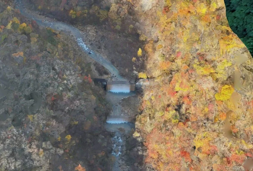 秋の渓谷を流れる川に、紅葉した木々に囲まれた2つの堰が見える空撮写真。