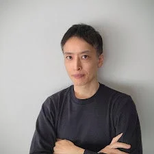 明るい灰色の壁を背景に、黒い長袖シャツを着て腕を組んだ日本人男性のポートレートです。