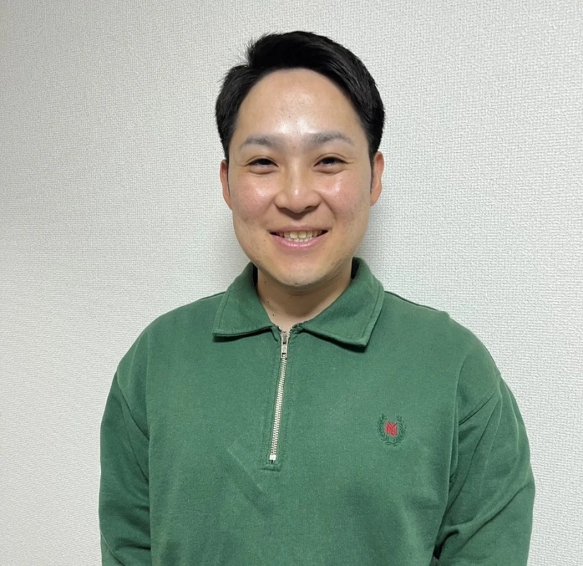 緑色の襟付きスウェットシャツを着たアジア人男性が、白い壁を背景に穏やかな笑顔を浮かべているポートレート写真