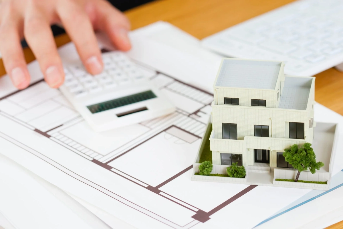 間取り図の上に置かれた住宅模型と、その奥で電卓を操作する手
