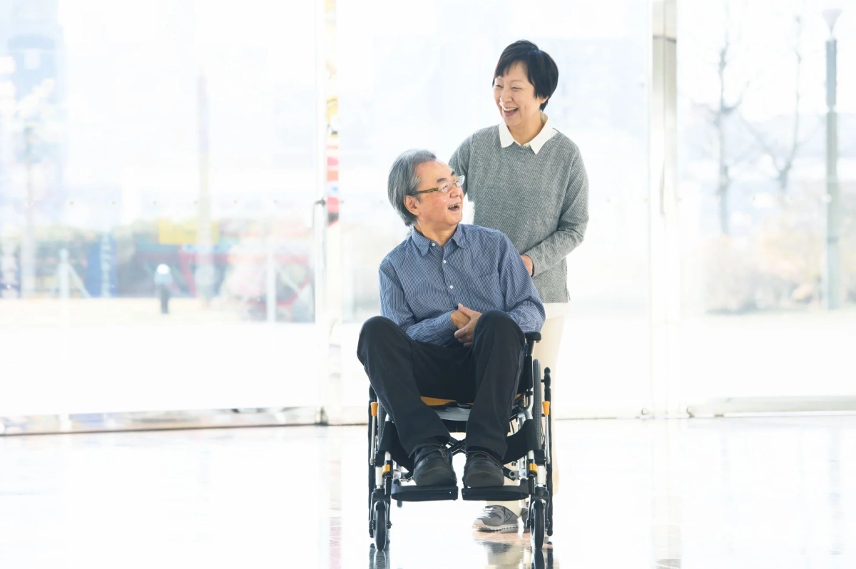 車椅子に座る高齢男性と、後ろで笑顔で話しかける高齢女性の様子