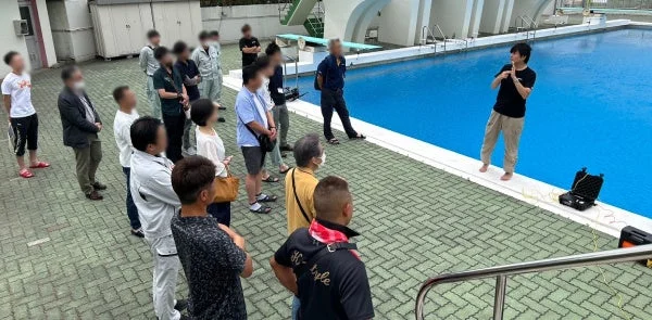 屋外プールサイドで、一人の男性が多数の人々に向けて何かを説明している場面