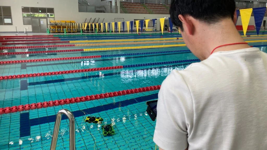 男性が競技用プールで2台の小型水中ロボットを操作・観察している様子