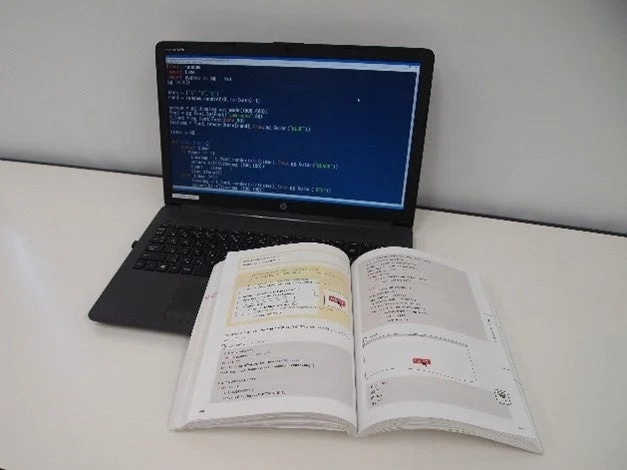 白い机の上にノートパソコンと開かれた教科書が置かれています