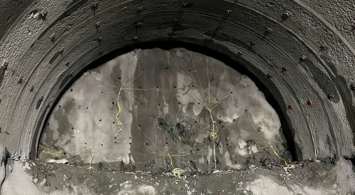 地下トンネルの建設現場の内部を写した画像。粗いコンクリートのアーチと、掘削中の岩盤に多数の穴と黄色いケーブルが設置されている様子