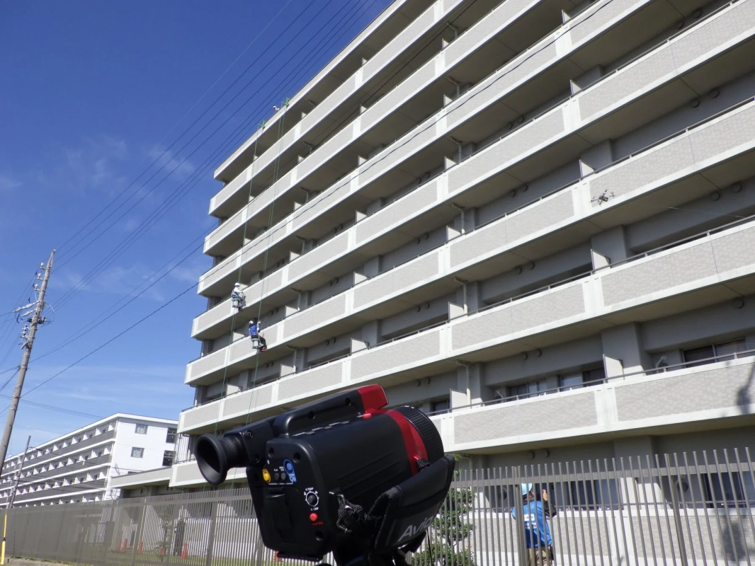 青空の下、高層マンションの外壁でロープアクセスによる作業員とドローンが確認できます