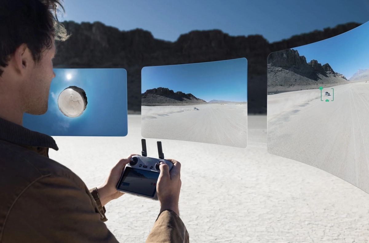 広大な砂漠でドローンを操作する人物が、リモートコントローラーと複数の仮想スクリーンを通じて、車両追跡や360度ビューを含む空撮映像を確認している様子。