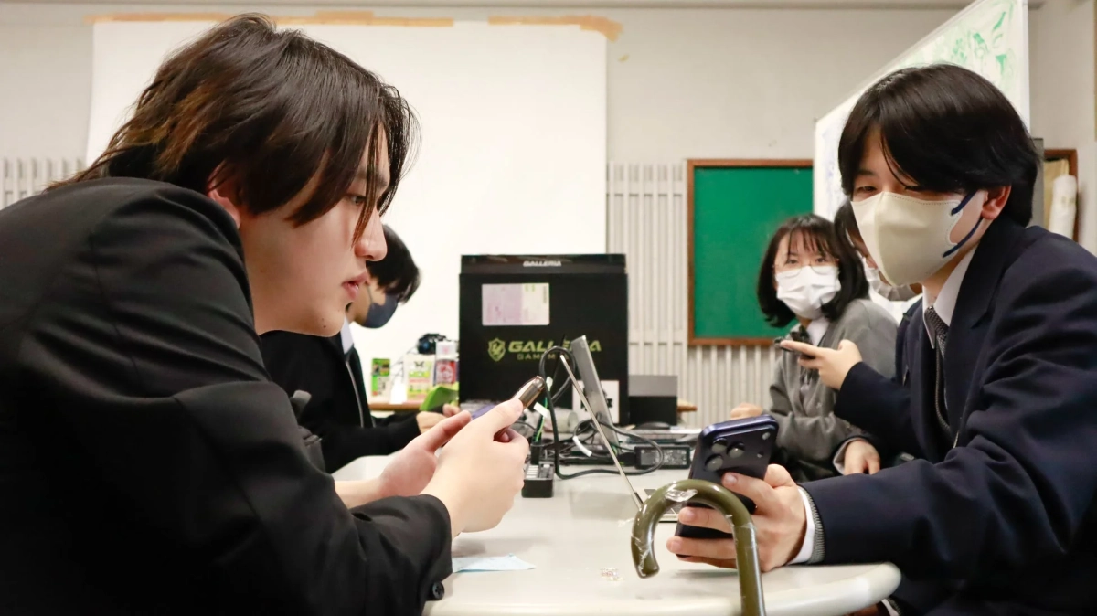 制服を着た若者たちがテーブルを囲み、スマートフォンを操作している様子。背景にはゲーミングPCが見え、部活動や学内での集まりのようだ。