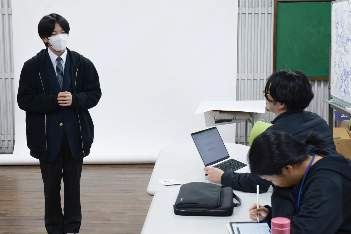 教室のような空間で、一人の人物が立って話しており、他の二人が座ってノートパソコンやタブレットを使用している様子です。