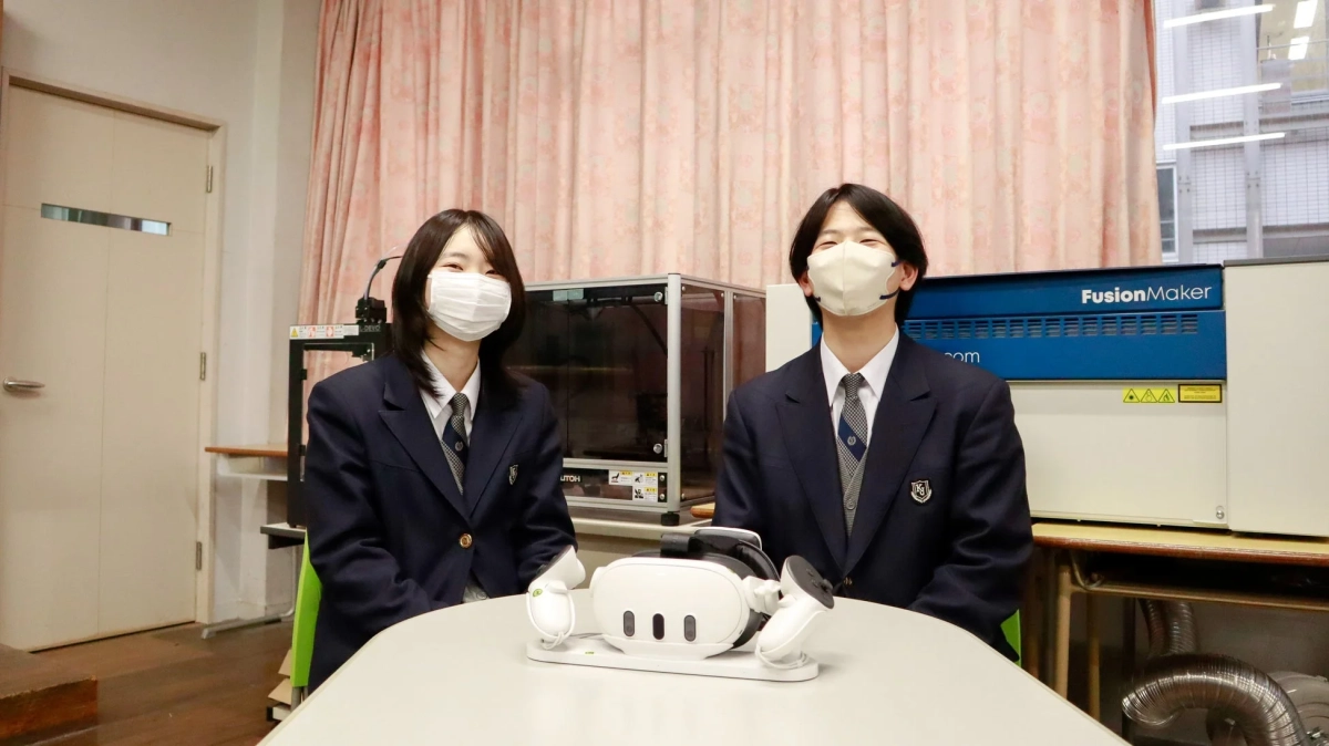 制服とマスク姿の男女学生が、VRヘッドセットを前に笑顔で座っています。学校のラボや教室のような場所で、最新技術に触れる教育活動の一場面を捉えた写真です。