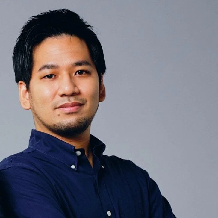 紺色のボタンダウンシャツを着たアジア系の男性が、シンプルなグレーの背景を背に、カメラをまっすぐ見ています。穏やかな表情のポートレートです。