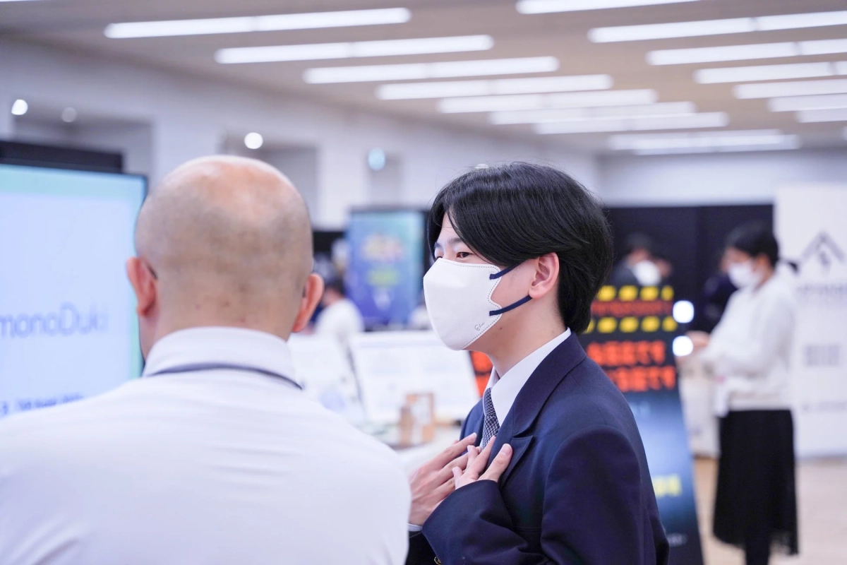 イベント会場で、スーツを着てマスクをした男性が、白いシャツを着た男性と話している様子。背景には他の参加者やディスプレイが見えます。