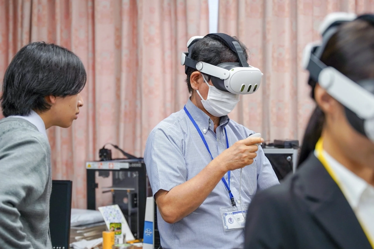 VRヘッドセットとマスクを着用した男性がコントローラーを操作し、VR体験をしている様子。若者が観察し、別の人物もVRヘッドセットを装着。仮想現実技術のデモンストレーションか研修の場面と推測される。
