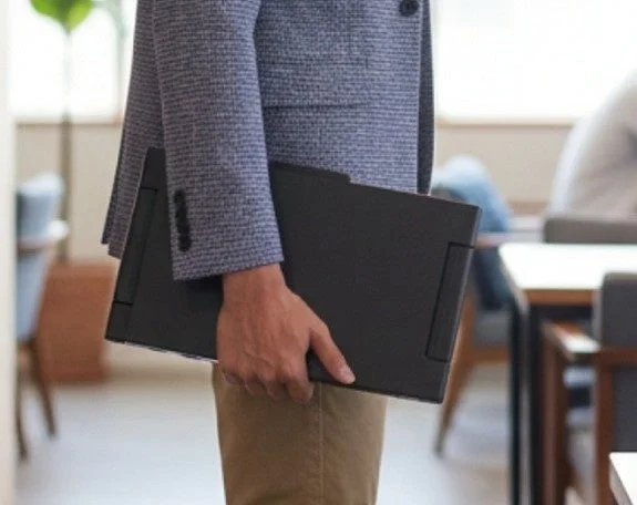 ビジネスシーンで、スーツのジャケットとチノパンを着用した人物がノートパソコンを抱えて歩いている様子。