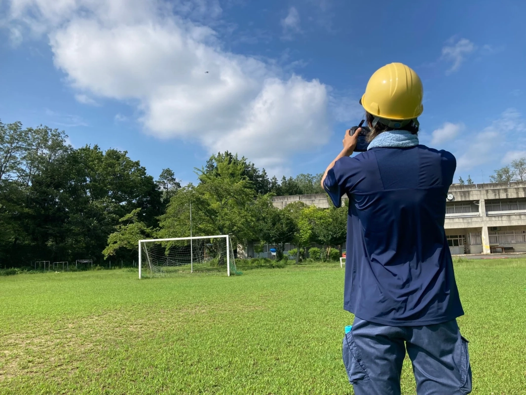 晴れた日の屋外のグラウンドで、黄色いヘルメットをかぶった人物がドローンを操作している様子