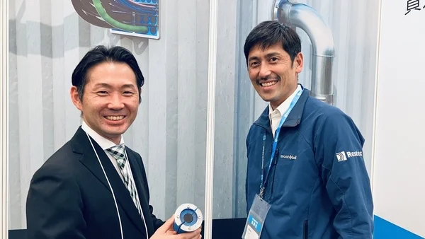 展示会のような場所で、笑顔の男性2人が並んで写っています。一人はスーツ姿で、もう一人はmont-bellのジャケットを着用しており、和やかなビジネスシーンを捉えています。