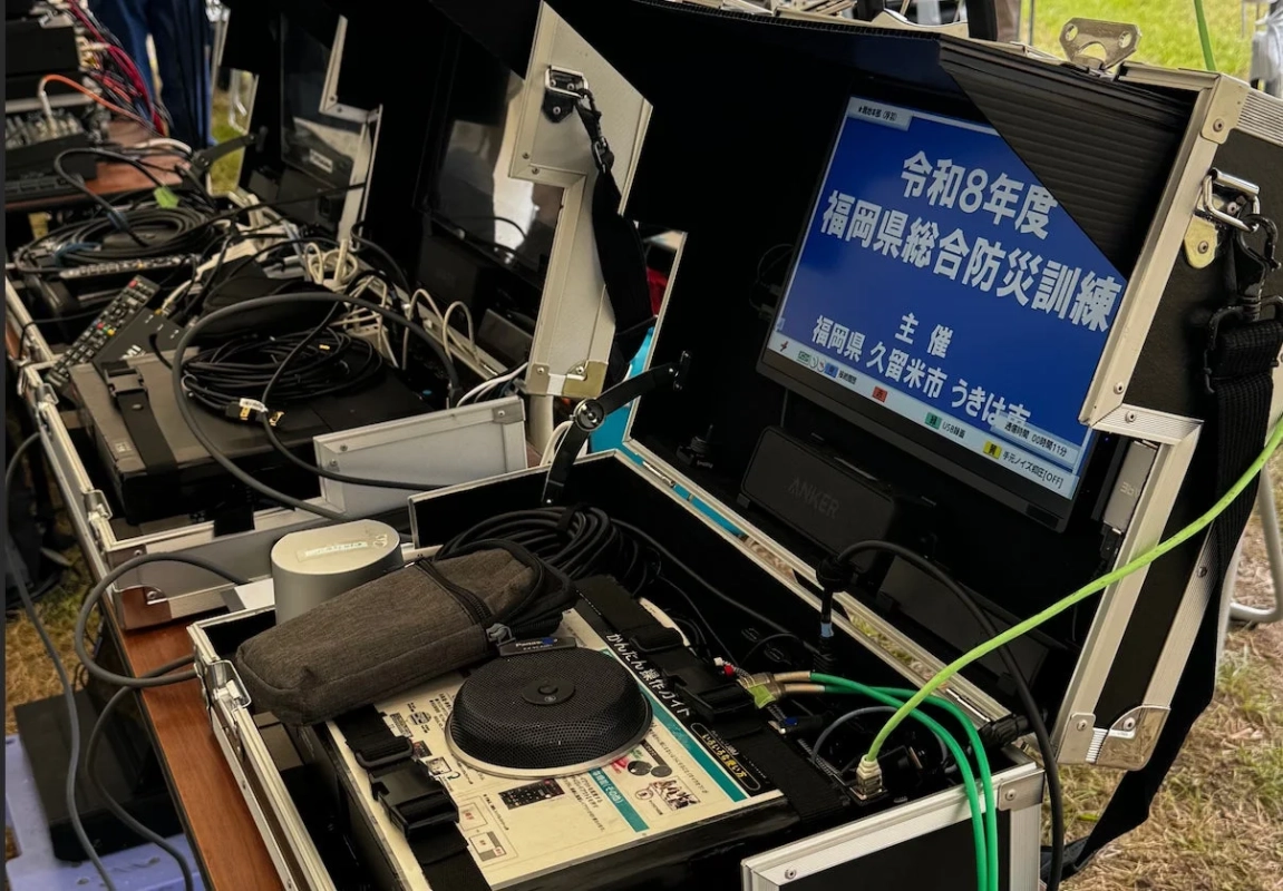 「令和8年度福岡県総合防災訓練」と表示されたモニターを中心に、多数のケーブルや電子機器が設置された屋外のイベント機材の様子。福岡県、久留米市、うきは市が主催する防災訓練の準備風景とみられる。
