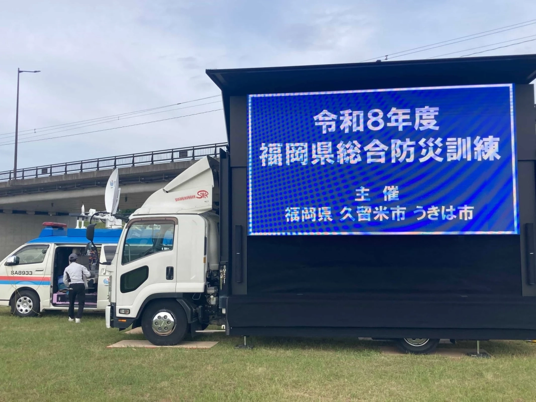令和8年度の福岡県総合防災訓練の様子を捉えた画像です。大型LEDトラックに訓練の開催情報が表示され、衛星通信設備を備えた緊急車両も待機しています。
