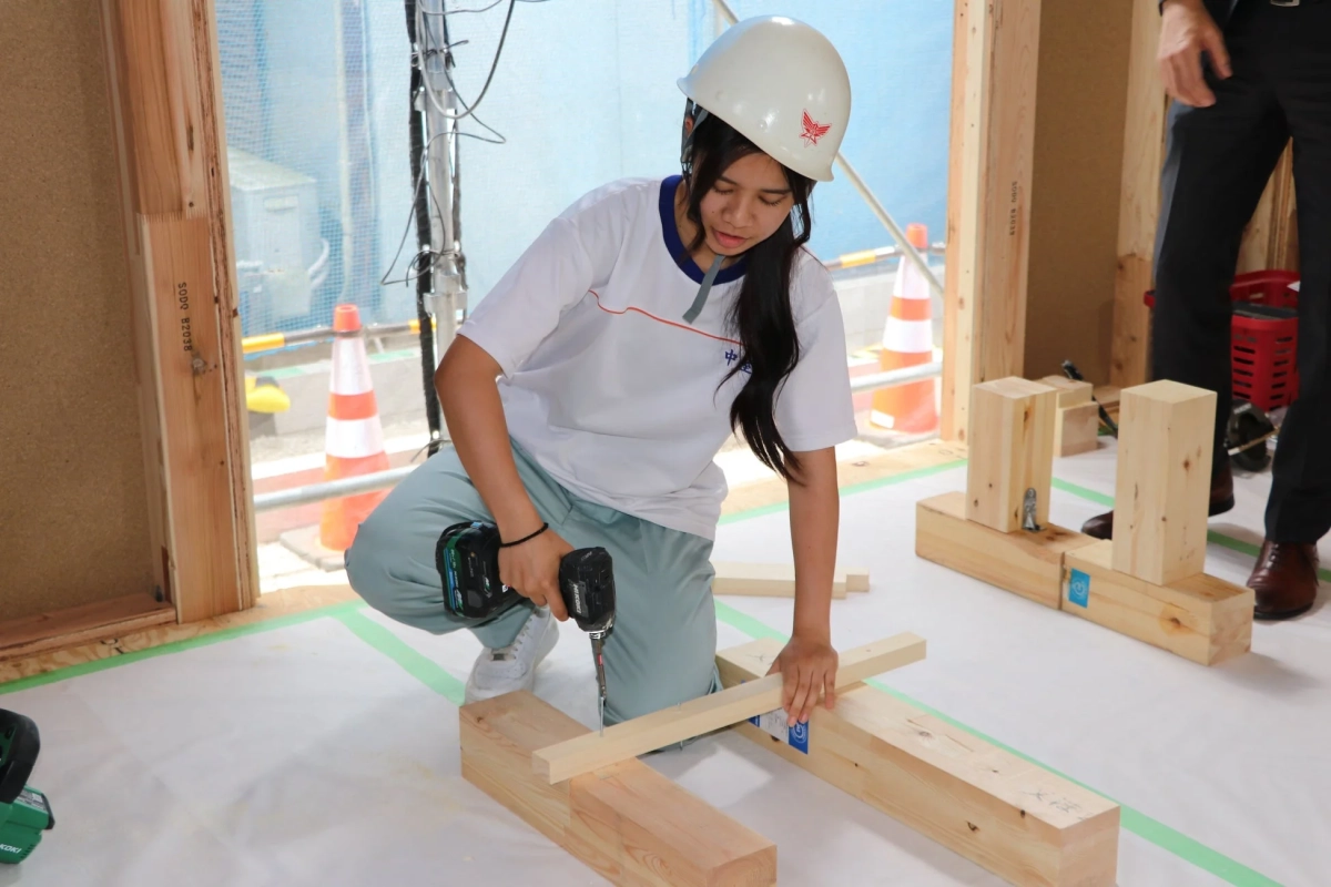 白いヘルメットをかぶった若い女性が、建設現場のような場所で電動ドリルを使って木材を組み立てる作業をしています