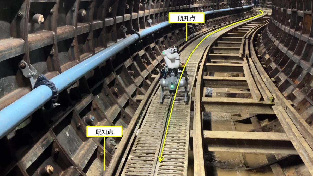 四足歩行ロボットが、青い配管が通る曲がりくねった地下トンネルの線路脇を歩いている様子