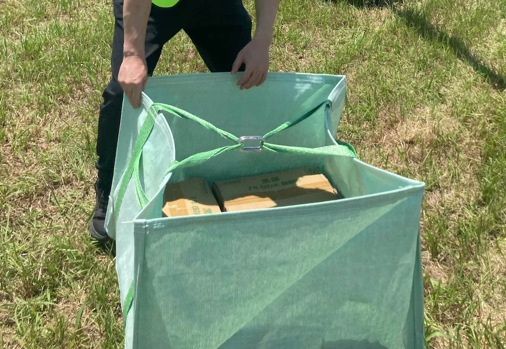 屋外の草地で、一人の人物が緑色の大きな袋を開けている様子です。袋の中にはいくつかの段ボール箱が入っており、そのうちの一つには「7% 消毒水」と書かれています。何かの運搬や作業中のようです。
