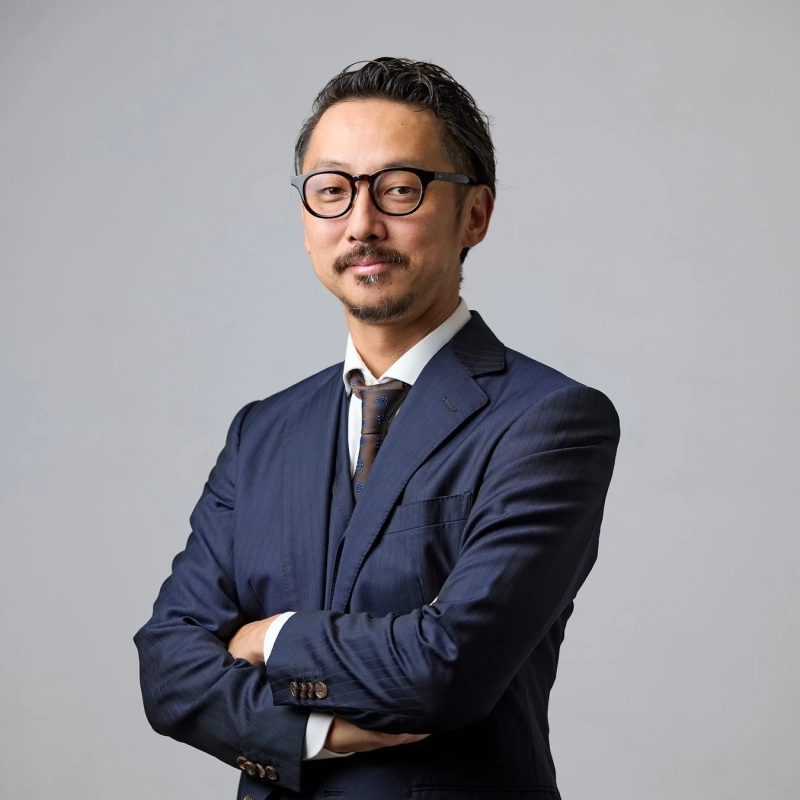 紺色のストライプスーツを着用し、眼鏡をかけたアジア人男性が腕組みをして立つポートレート。自信に満ちたプロフェッショナルな雰囲気です。