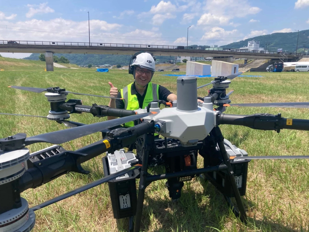 青空の下、広々とした草地で、安全ベストとヘルメットを着用した男性が大型ドローンの隣でサムズアップしています