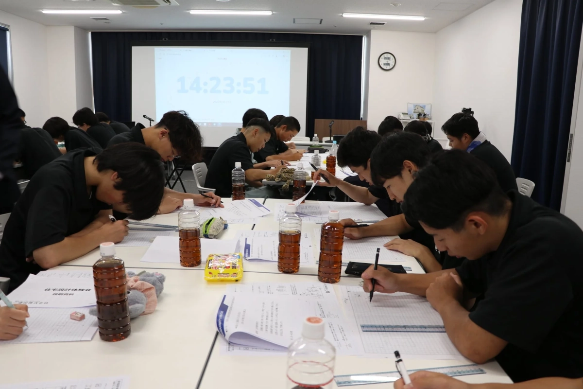 多くの若者が教室で机に向かい、真剣な表情で書類に書き込みや描画を行っています