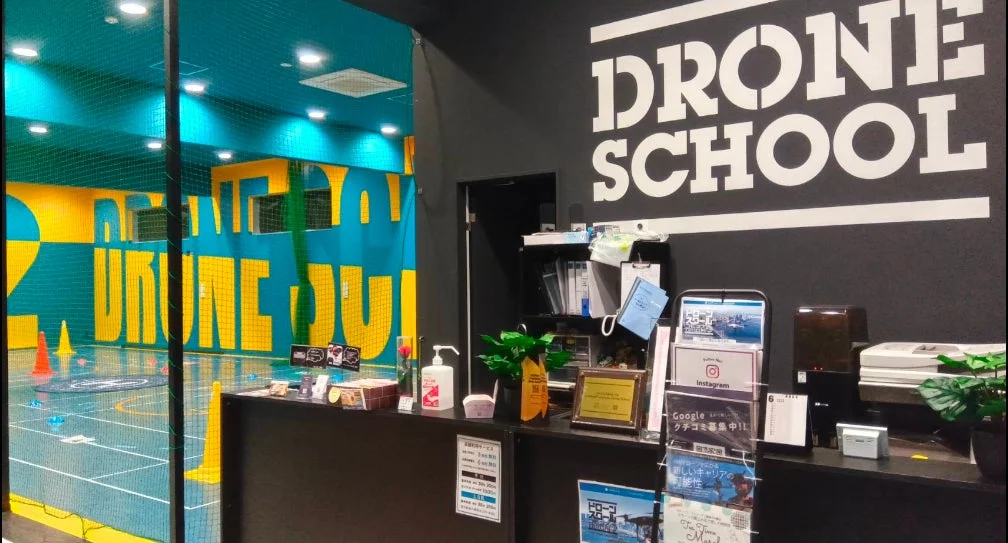 ドローンスクールの受付と室内練習場です。壁には「DRONE SCHOOL」の文字が大きく表示され、受付には関連資料や消毒液があります。