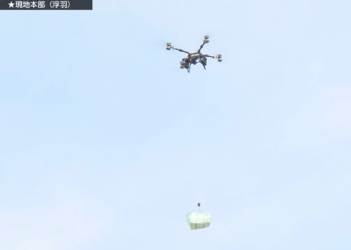 青空を背景にドローンが飛行しており、その下には荷物が投下され降下している様子が捉えられています。ドローンによる空中輸送や物資の配達を示唆するシーンです。