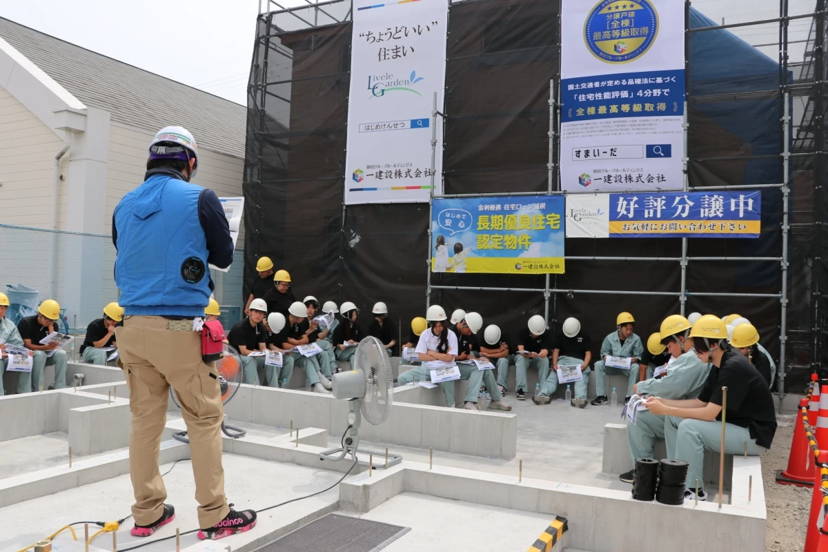 建設現場で、作業員たちが建物の基礎工事現場の周りに座り、監督らしき人物からの説明を聞いている様子